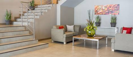 A modern living room with a staircase, grey sofas, a coffee table, and potted plants.