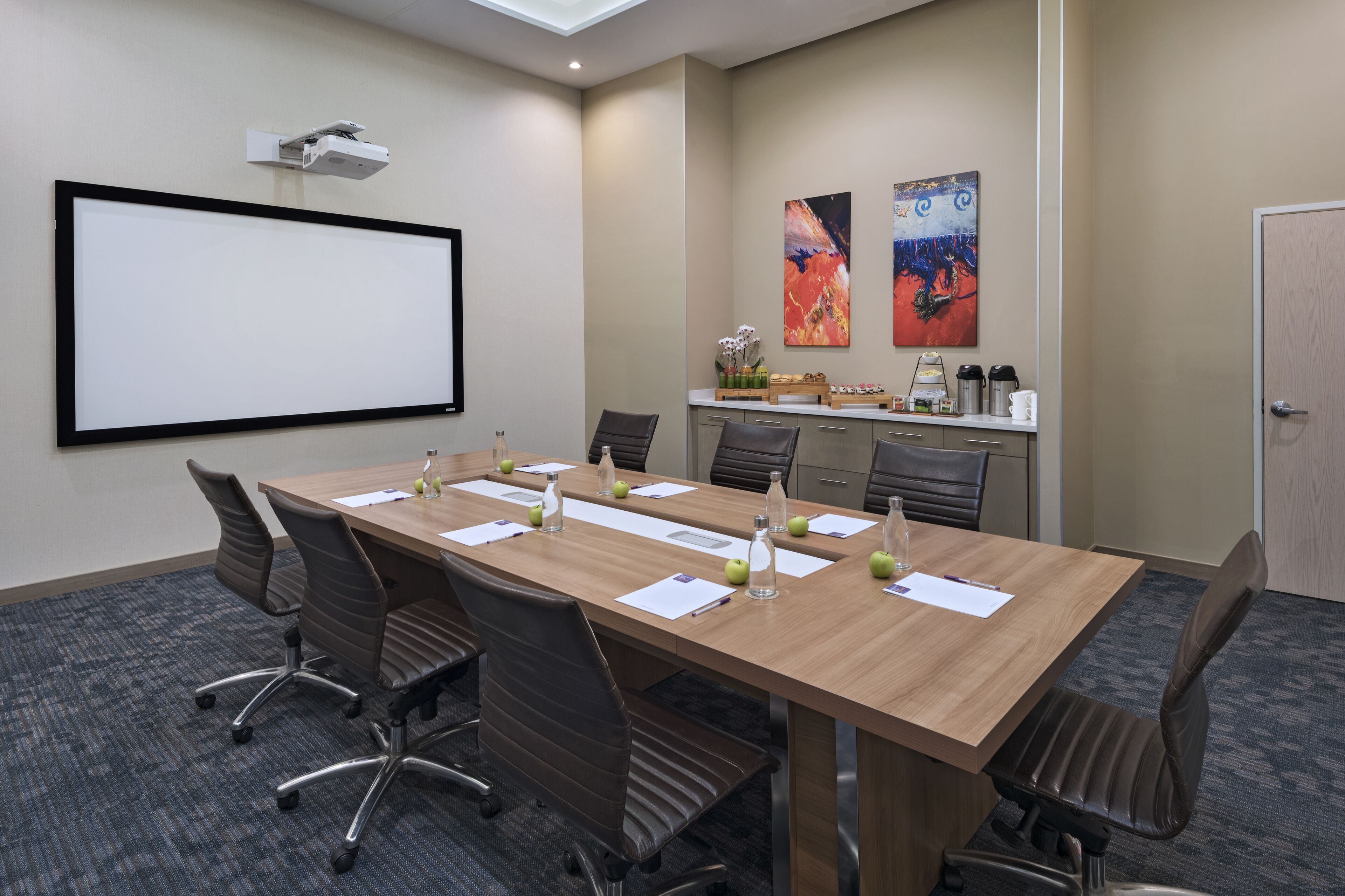 A conference room with a long table, chairs, a projector screen, and a coffee station.