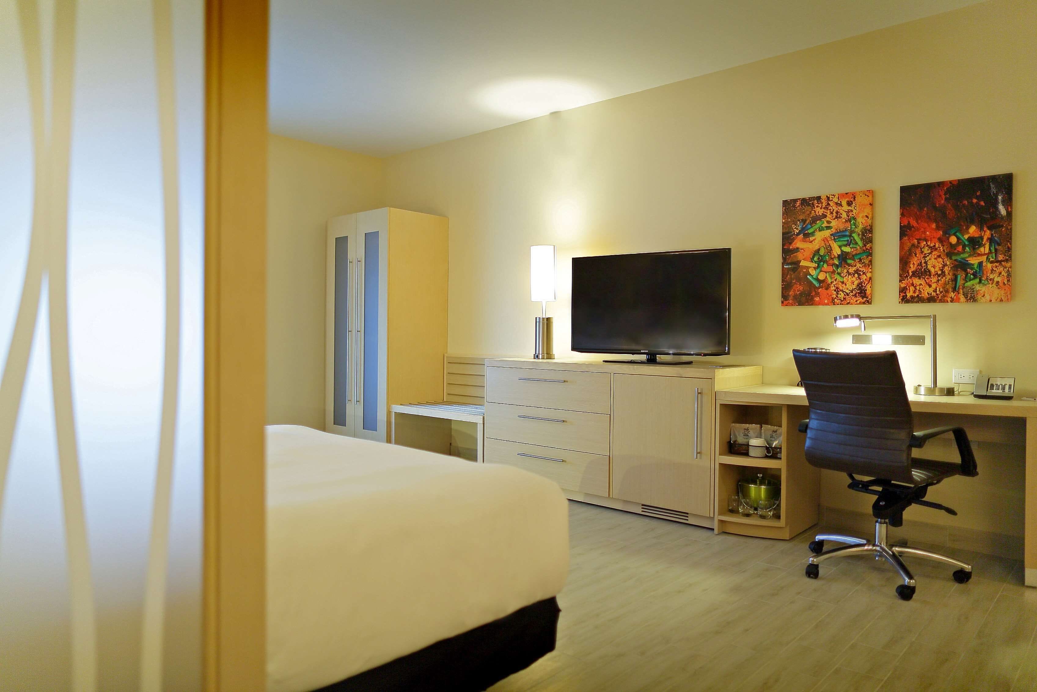 Una habitación de hotel con cama, escritorio, silla, televisor y cuadros en la pared.