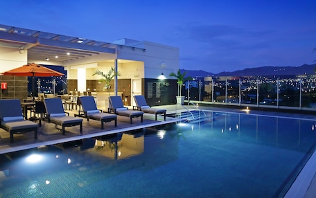 A rooftop infinity pool with lounge chairs and a dining area, overlooking a cityscape at dusk.