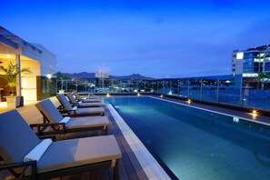 Un área de piscina en la azotea con sillones de descanso y vistas a la ciudad y montañas al fondo.
