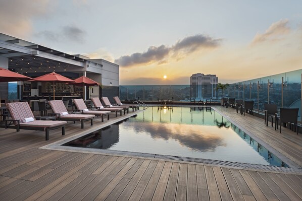 Área de piscina en la azotea con sillones de descanso y sombrillas, un bar y vistas a la ciudad al atardecer.