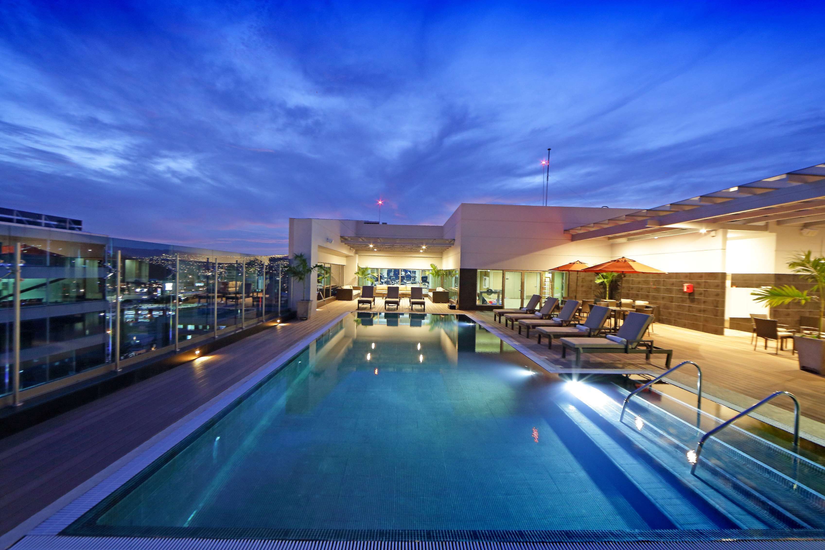 A modern indoor swimming pool with lounge chairs and umbrellas, surrounded by glass walls and a city view.