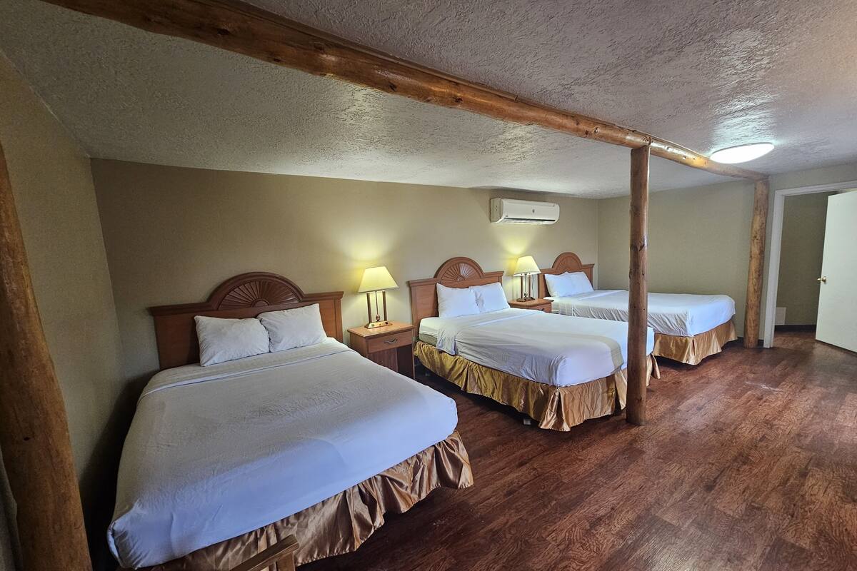 A hotel room with three single beds, wooden headboards, bedside tables with lamps, and an air conditioning unit on the wall.