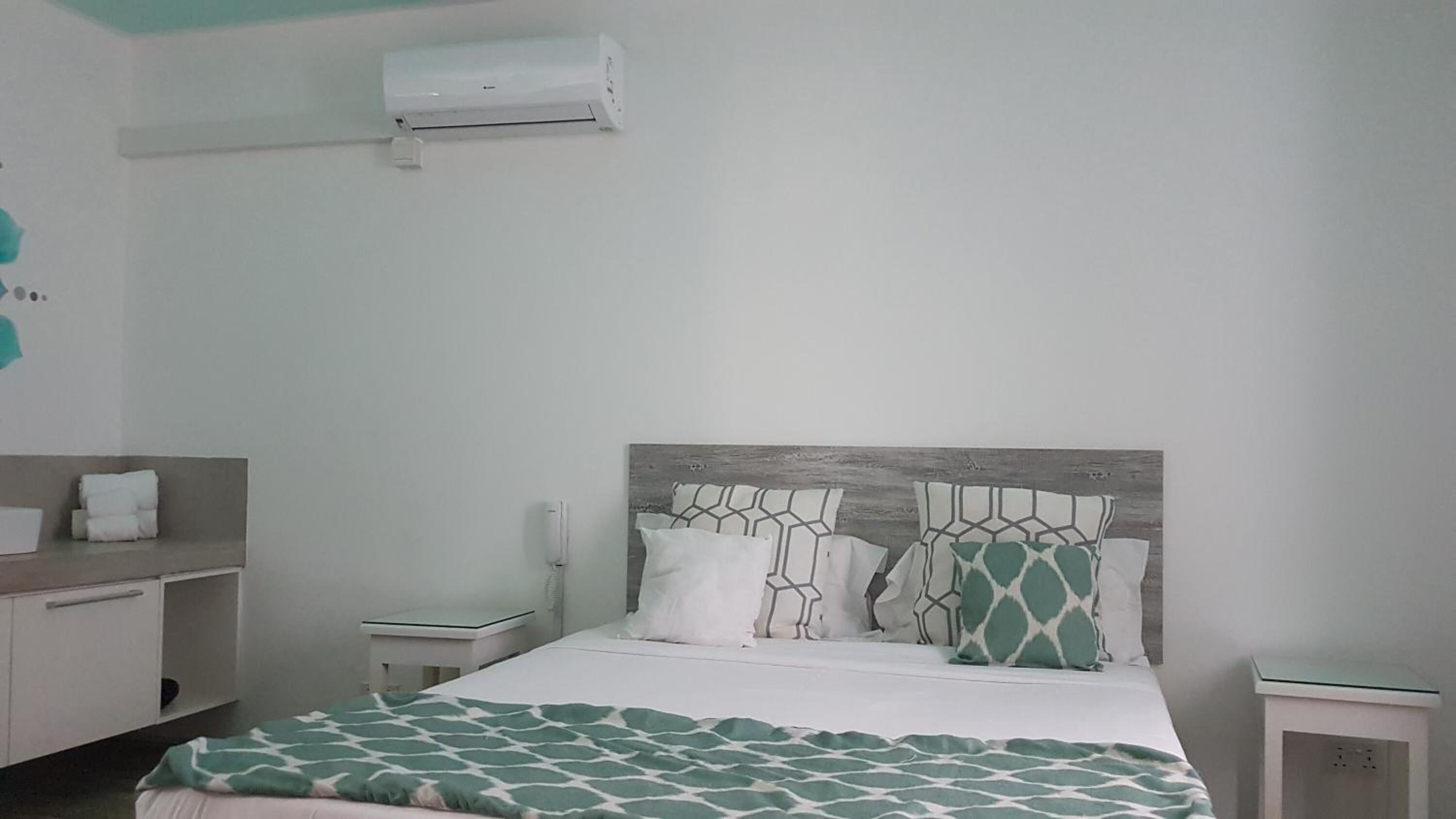 A hotel room with a bed, a bedside table, a nightstand, a wall-mounted air conditioner, and a bathroom counter with towels.