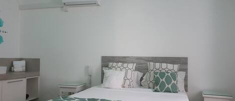 A hotel room with a bed, a bedside table, a nightstand, a wall-mounted air conditioner, and a bathroom counter with towels.