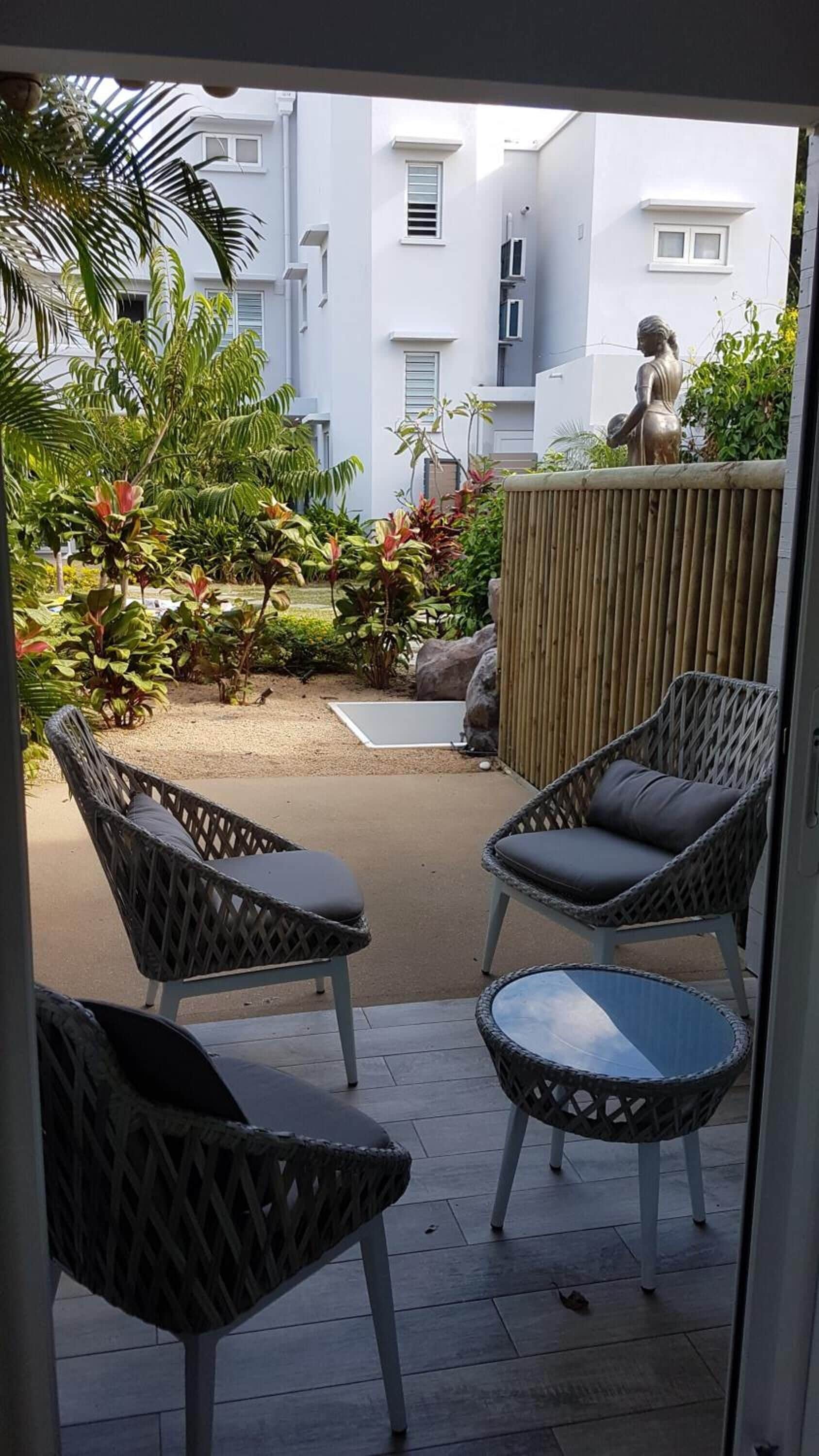 A patio area with wicker chairs and a small table, surrounded by lush greenery and a view of residential buildings.