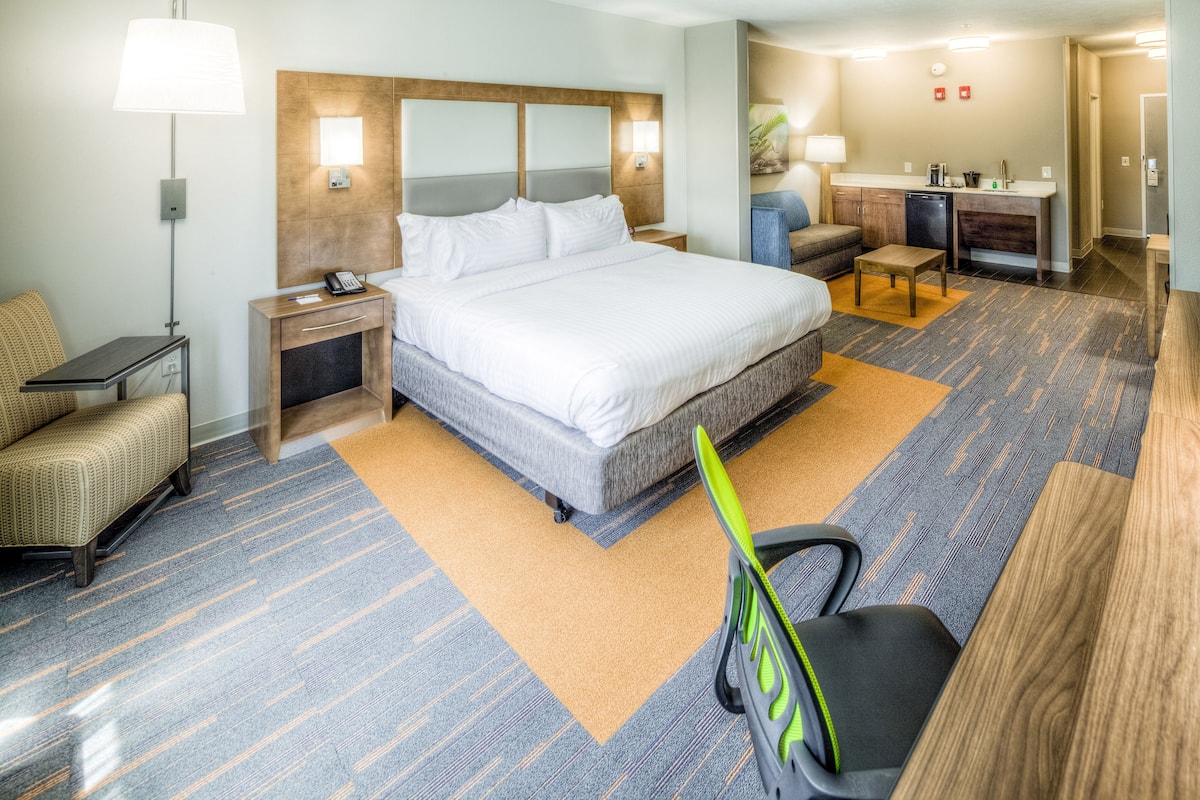 A hotel room with a large bed, a desk, a chair, a nightstand, a lamp, and a kitchenette.