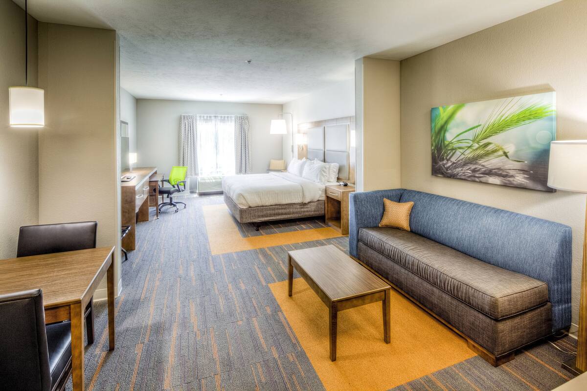 A hotel room with a bed, a sofa, a desk with a chair, and a painting on the wall.