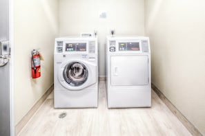Two front-loading commercial washers in a narrow hallway with a fire extinguisher mounted on the wall. - Holiday Inn Express & Suites Cleveland West - Westlake by IHG (Westlake)
