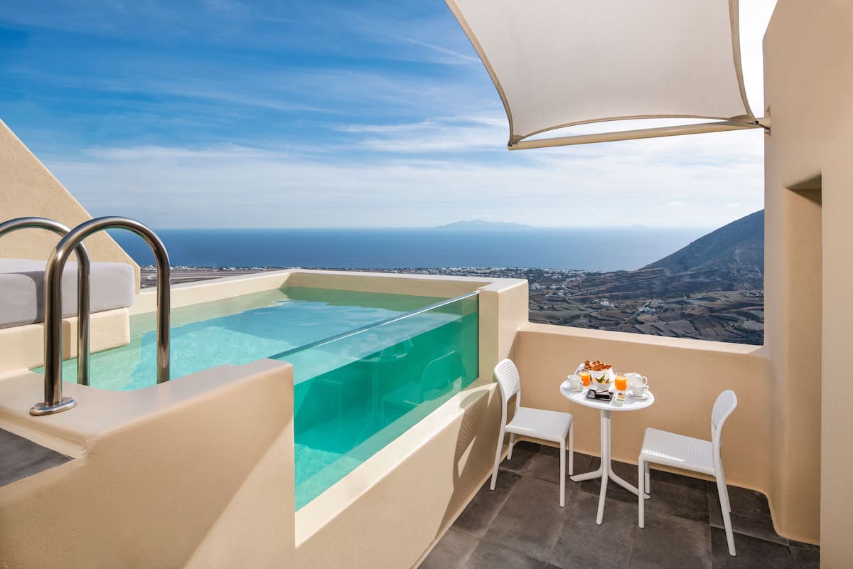 Un balcón con piscina infinita de cristal, sillas blancas y una mesa con alimentos para el desayuno.