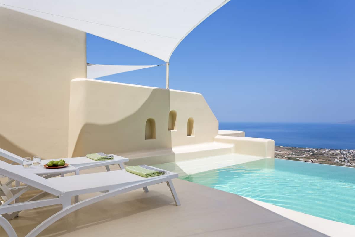 Un área junto a la piscina con sillas de descanso blancas, una mesa con una fuente de fruta y vistas al mar.