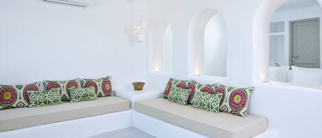 A modern, minimalist room with white walls, a built-in bench with patterned cushions, a small decorative bowl, and a ceiling-mounted air conditioner.