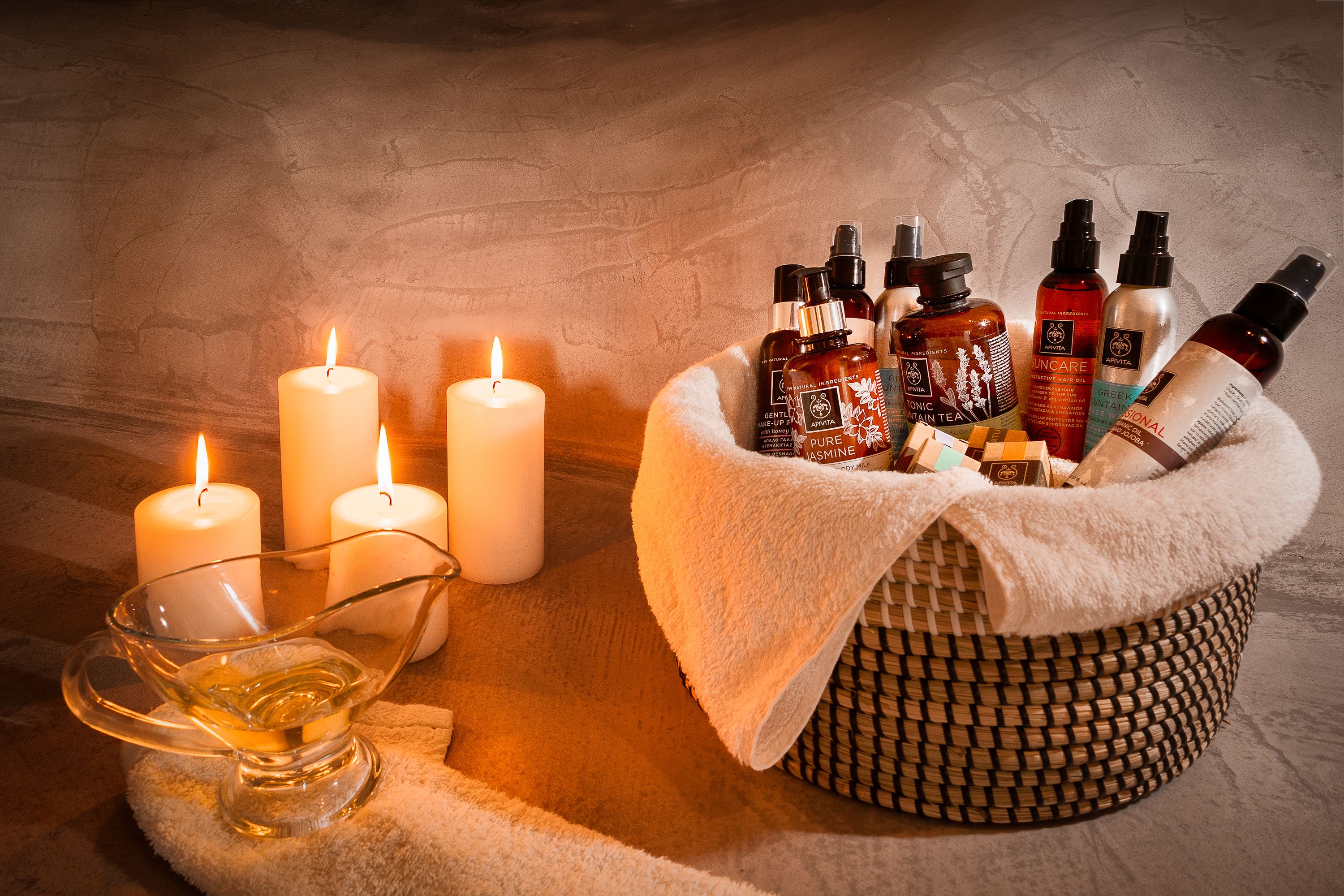 A candle-lit setting with a glass bowl of oil, a basket of skincare products, and lit candles.