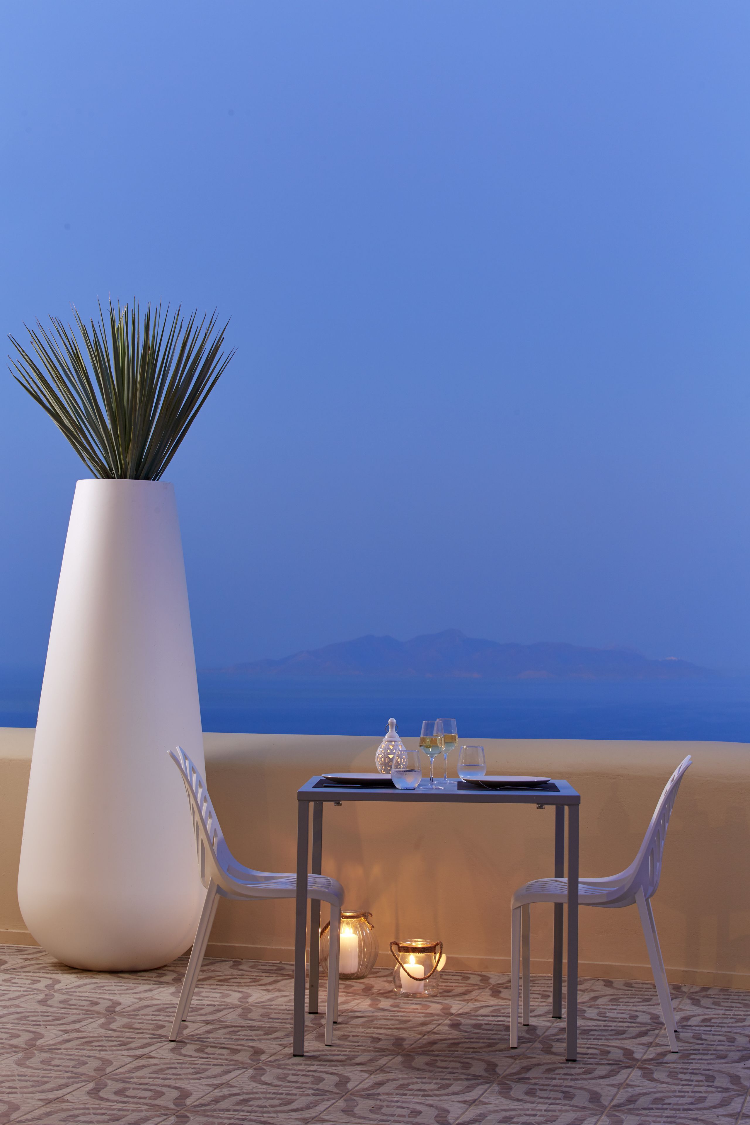 A dining table with two chairs, a large potted plant, and a candle on a terrace with a view of the sea and mountains.
