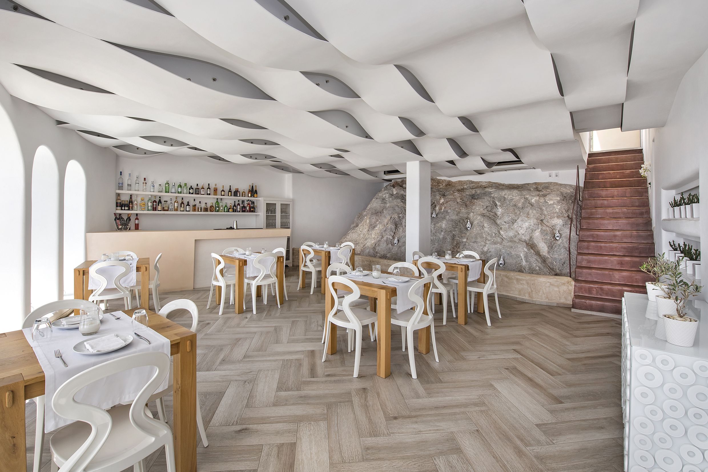Un restaurante moderno con mesas de madera y sillas blancas, suelo con diseño de espiga y una zona de bar con pared de piedra y escalera.