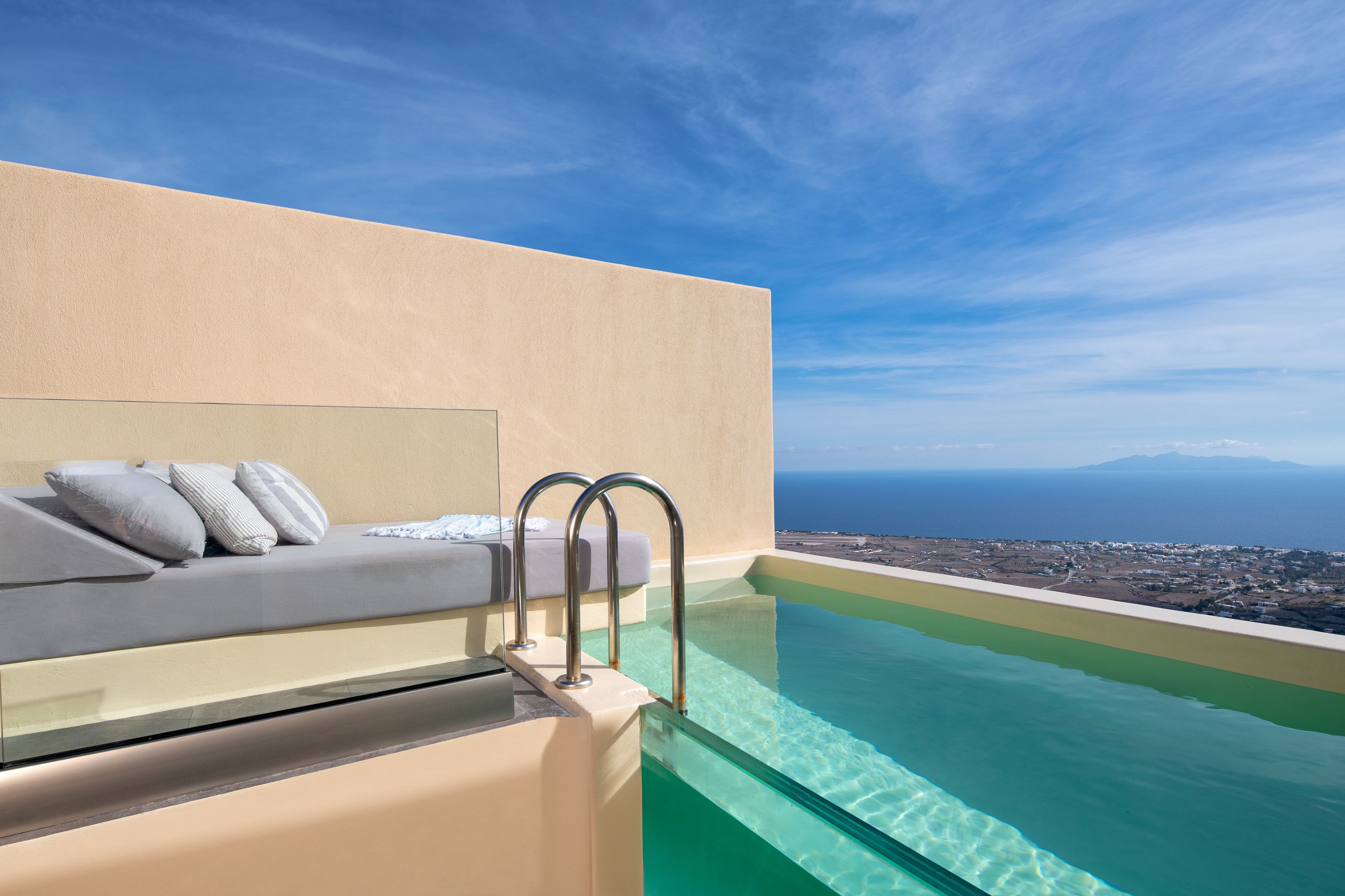 A modern infinity pool with clear water, a beige wall, and a sofa with pillows.