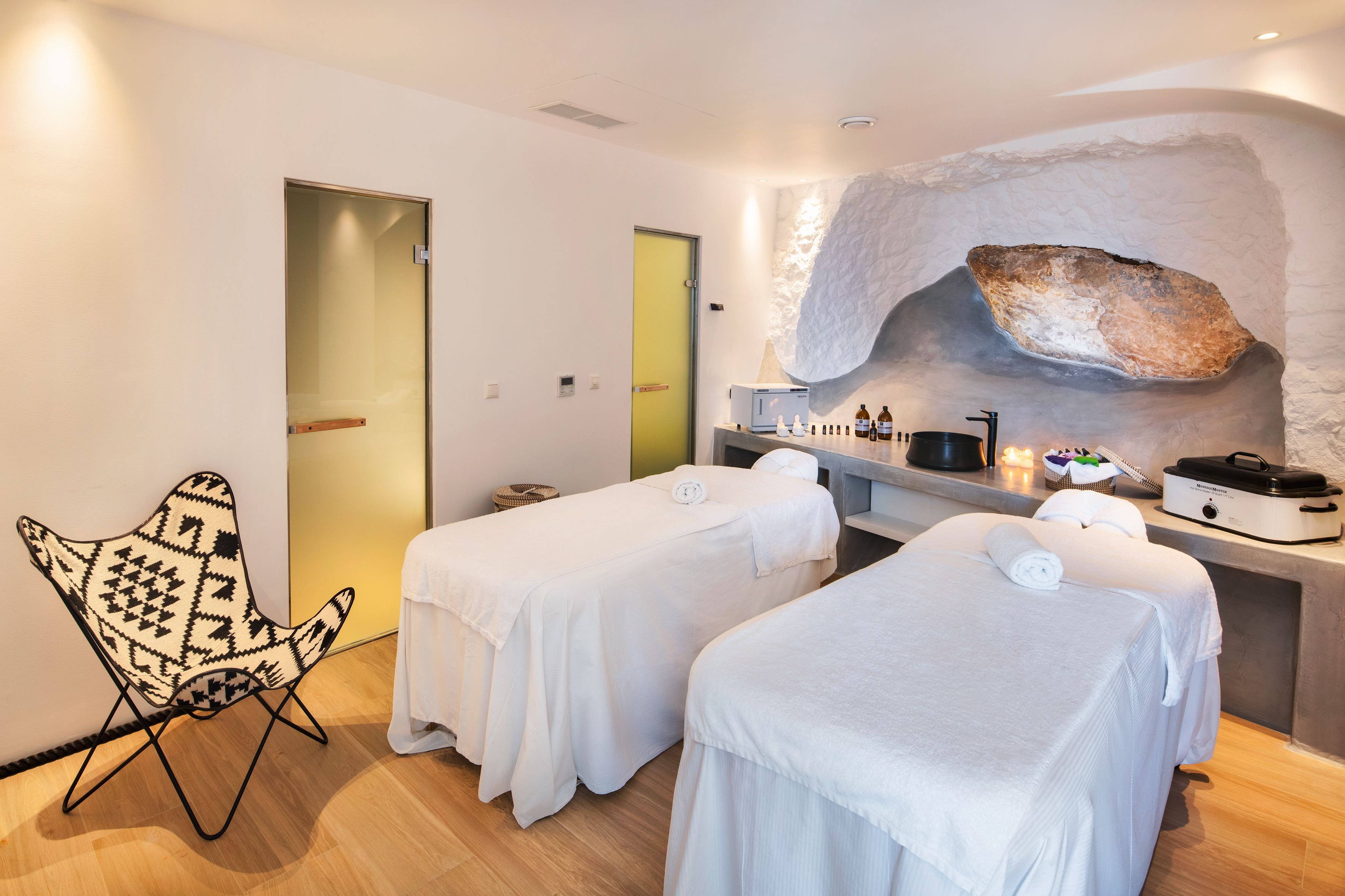 A spa room with two massage beds, a decorative chair, and a stone wall feature.