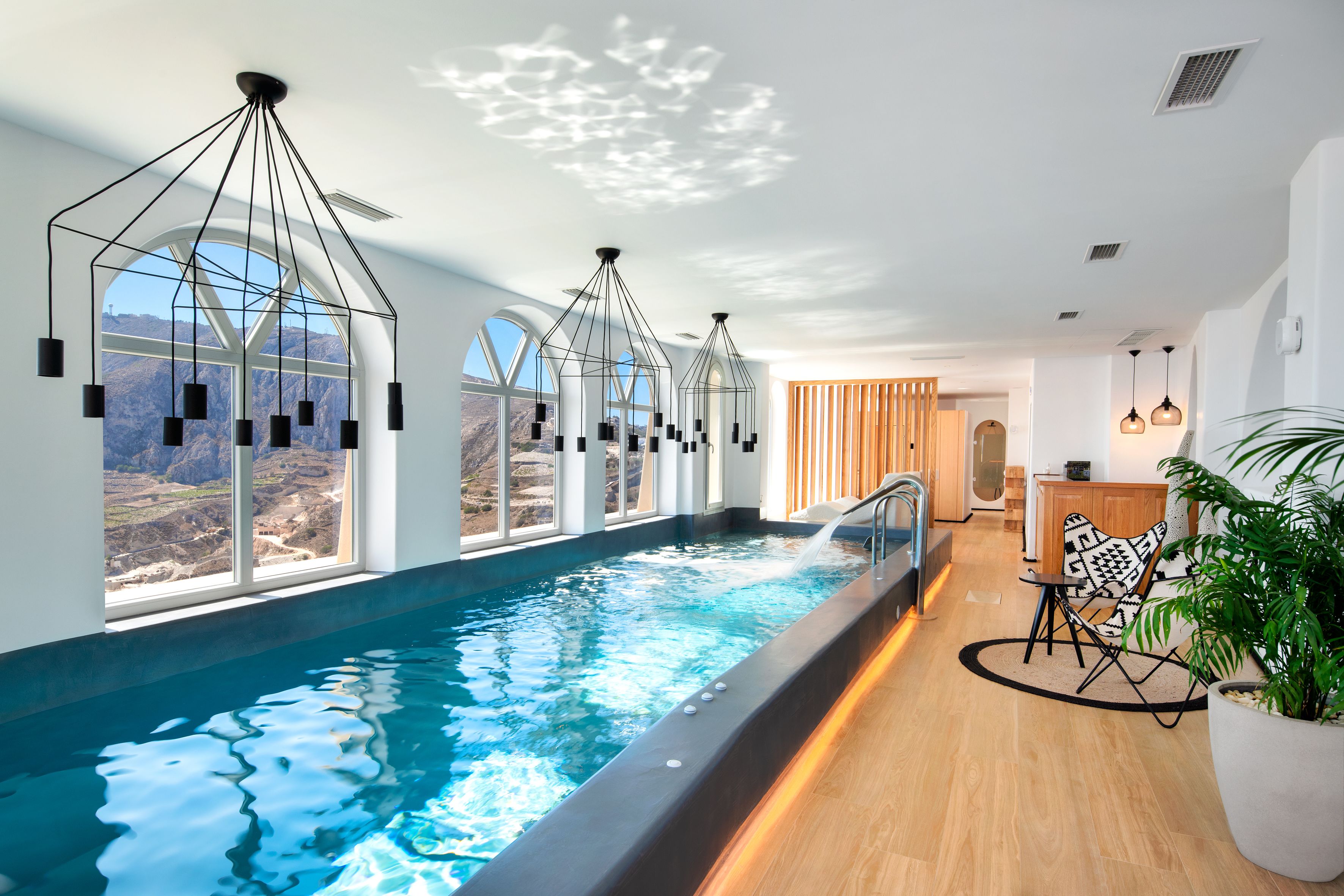 Une piscine intérieure moderne offrant une vue dégagée sur les montagnes, avec un plancher en bois et un espace détente agrémenté d’une grande plante en pot.