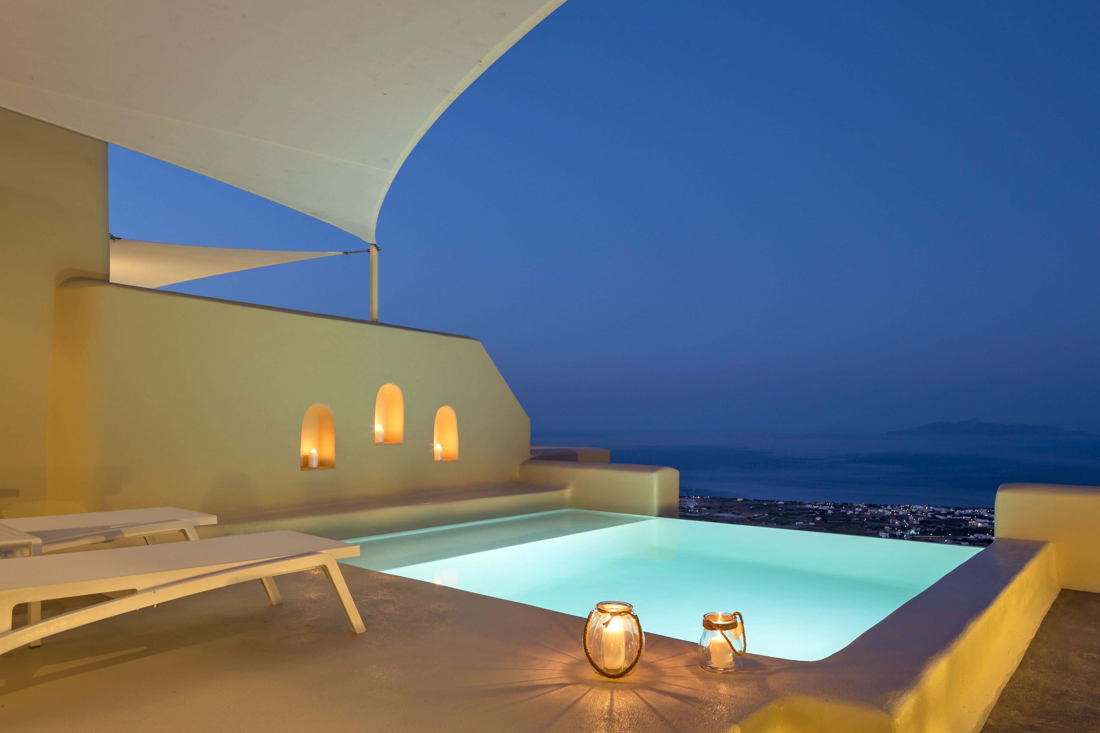 Piscina infinita en la azotea con sillones de descanso y velas, con vistas a un paisaje urbano al atardecer.