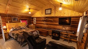 A log cabin room with a bed, two leather chairs, a fireplace, a television, and wooden walls.