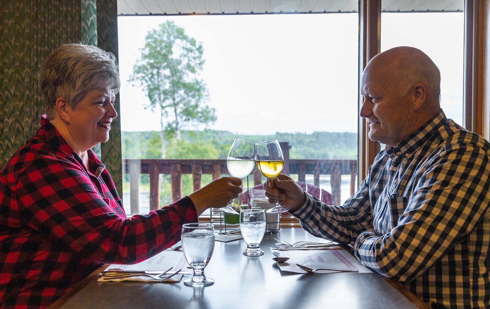 Deux personnes trinquent avec des verres de vin, assises à une table d’où l’on aperçoit des arbres et une terrasse en bois.