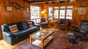 A cozy cabin interior with a leather sofa, two armchairs, a coffee table, and a large window offering a view of the outdoors.
