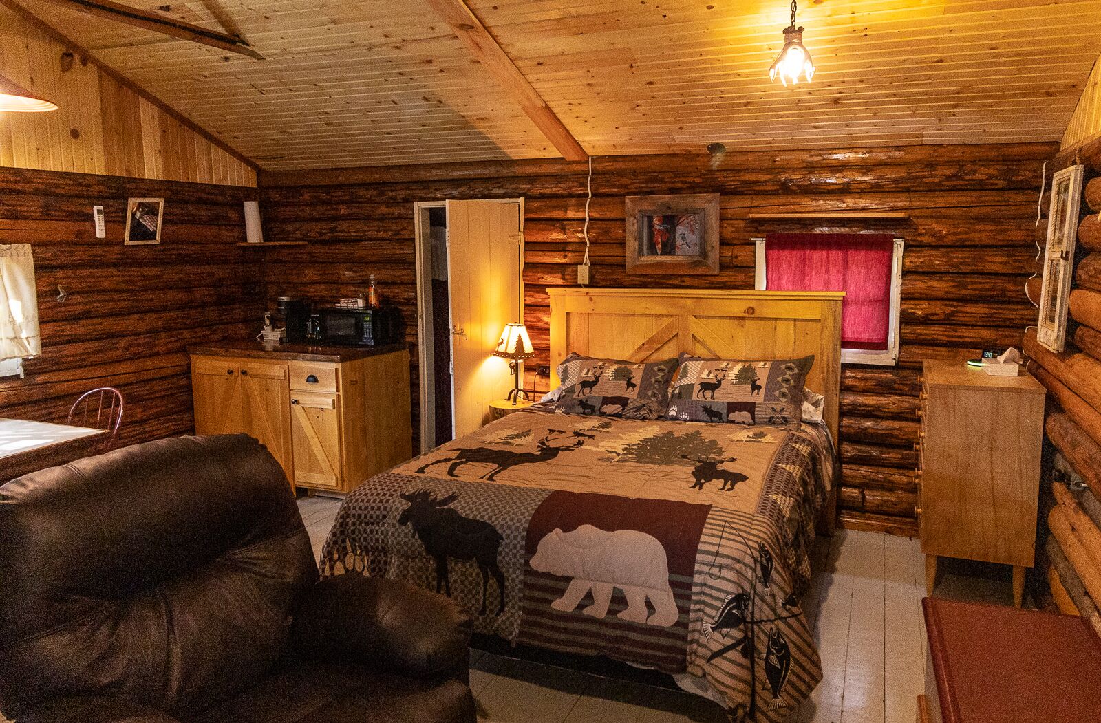 Acogedora habitación tipo cabaña de madera con cama, escritorio, silla y pequeña cocina.