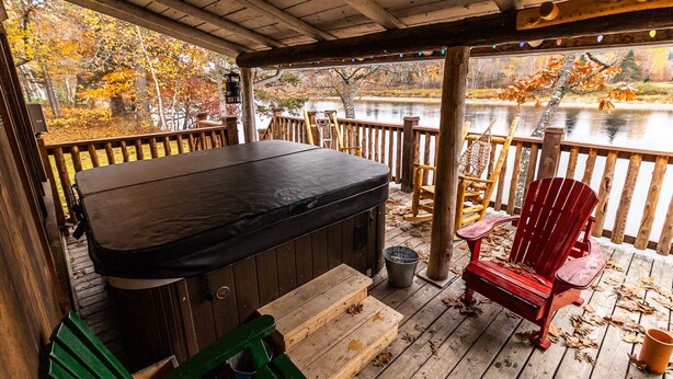 A wooden deck with a hot tub, red Adirondack chairs, and a view of a lake surrounded by autumn foliage.