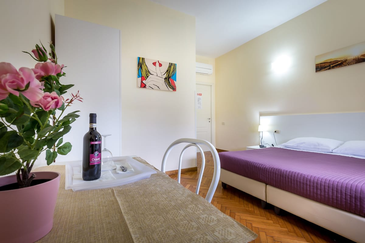 A hotel room with a bed, a chair, a table with a bottle and glass, a potted plant, and artwork on the walls.