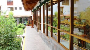 Un pasillo exterior cubierto con ventanas amplias y vistas a un patio con plantas y un edificio al fondo.