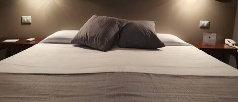 A neatly made bed with two pillows, a bedside table with a phone and a small item, and wall-mounted lighting.