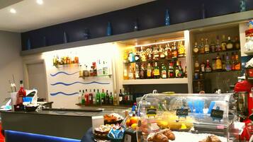 Zona bar con scaffali pieni di bottiglie di liquori, un bancone con pasticcini e una macchina da caffè, e una parete decorata con linee blu ondulate.