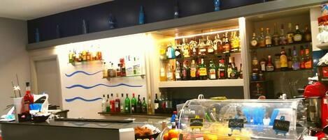 Zona de bar con estantes llenos de botellas de licor, una barra con pasteles y una máquina de café, y una pared decorativa con líneas azules onduladas.