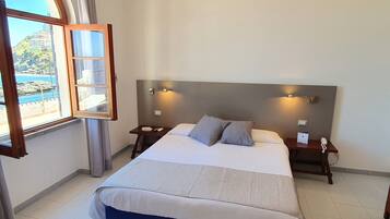 Een slaapkamer met een bed, nachtkastjes, een lamp en een raam met uitzicht op een kustlandschap.