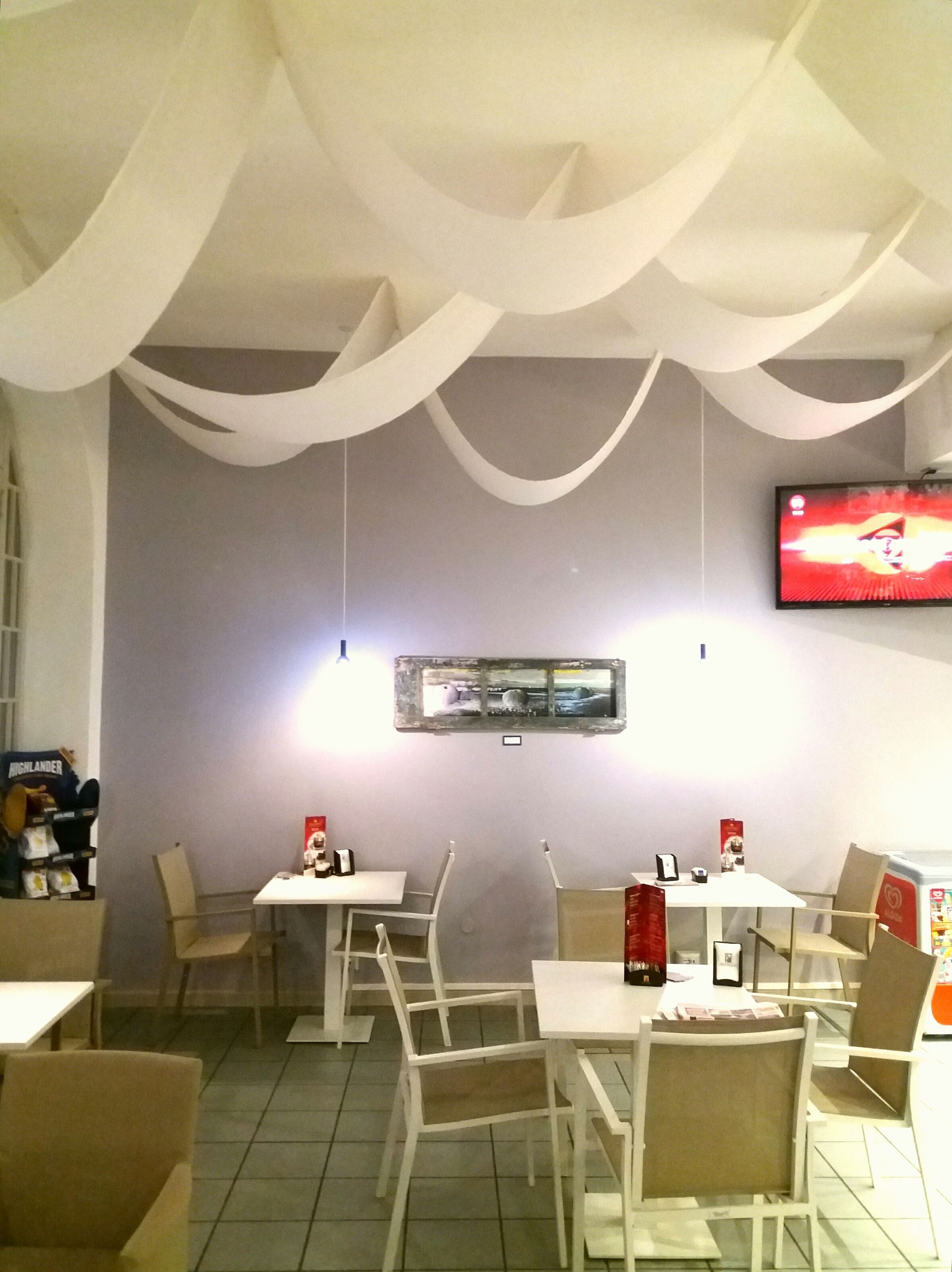Ein Restaurantinnenraum mit weißen Stühlen und Tischen, einem an der Wand befestigten Flachbildfernseher und einer dekorativen Decke.
