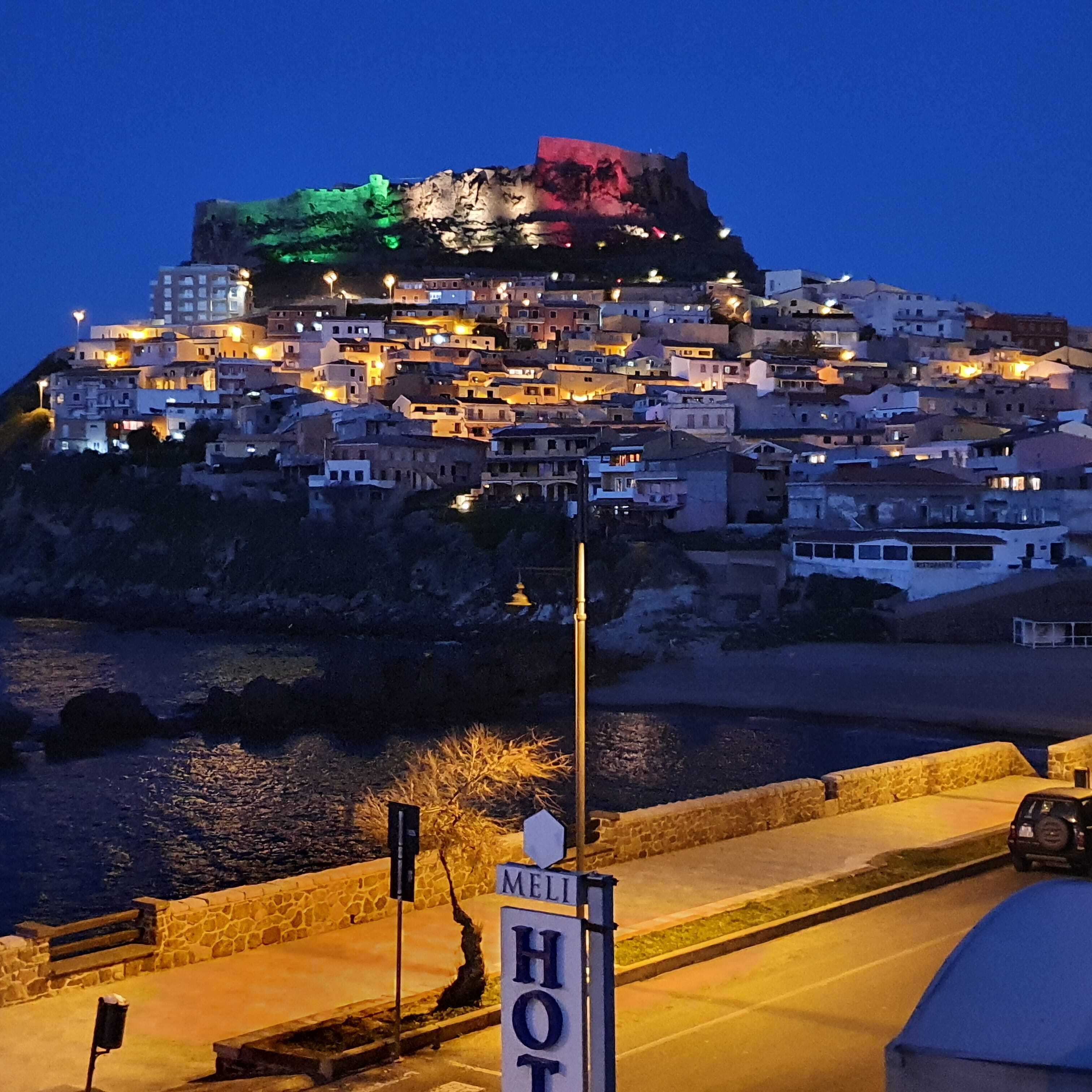 Una località costiera illuminata di notte, con edifici illuminati. Un cartello indica un albergo chiamato "HOTEL MELLI".