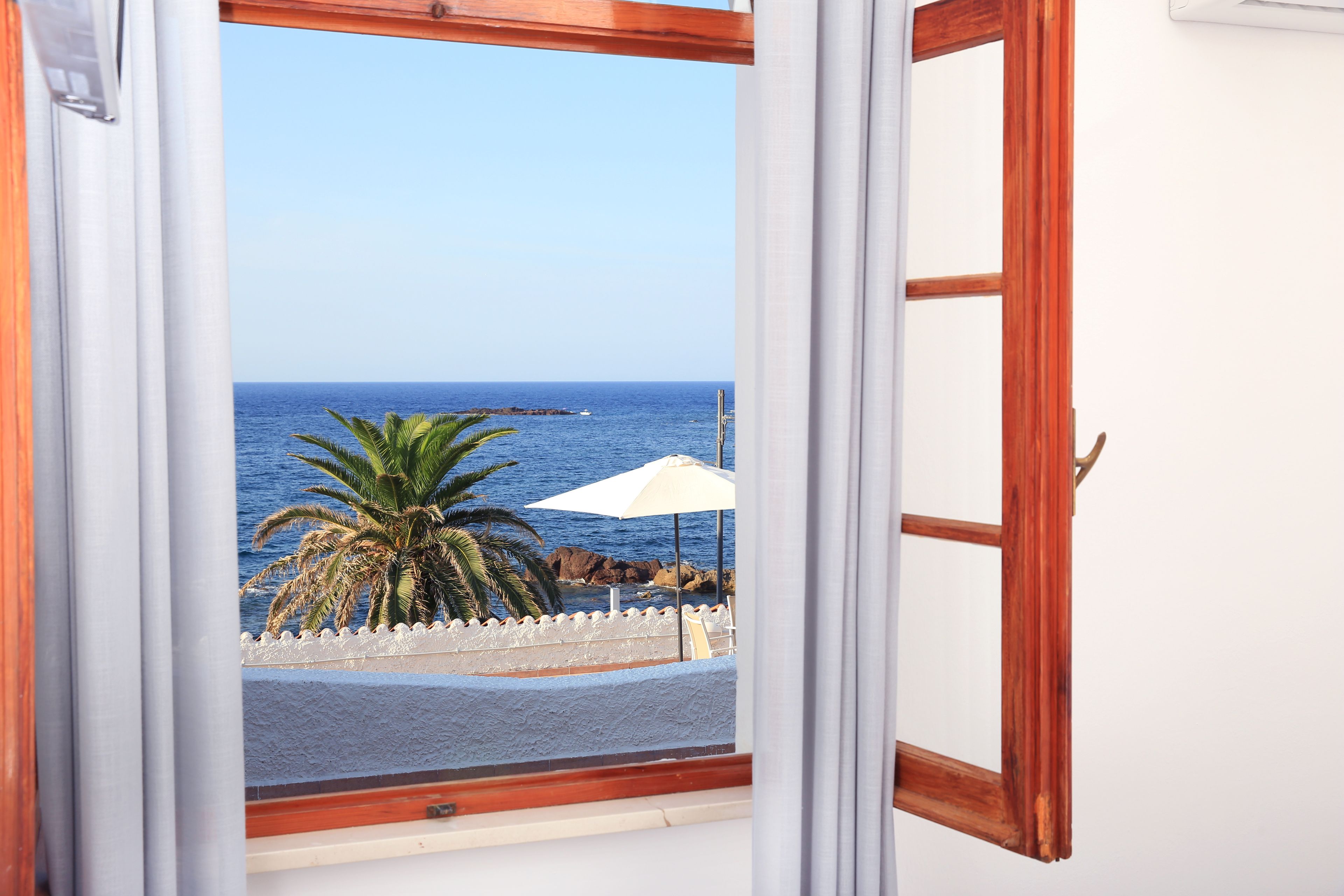 Vista desde una ventana con marco de madera y cortinas blancas, que da a una playa con una palmera y una sombrilla blanca.