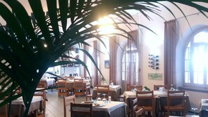 Un restaurante con varias mesas y sillas, ventanas amplias y plantas en macetas.