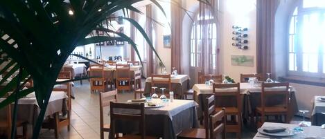A restaurant with multiple tables and chairs, large windows, and potted plants.