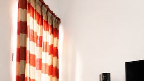 Una habitación con un sillón gris, un calentador negro alto, una cortina a rayas roja y beige, y un televisor sobre un mueble de madera.
