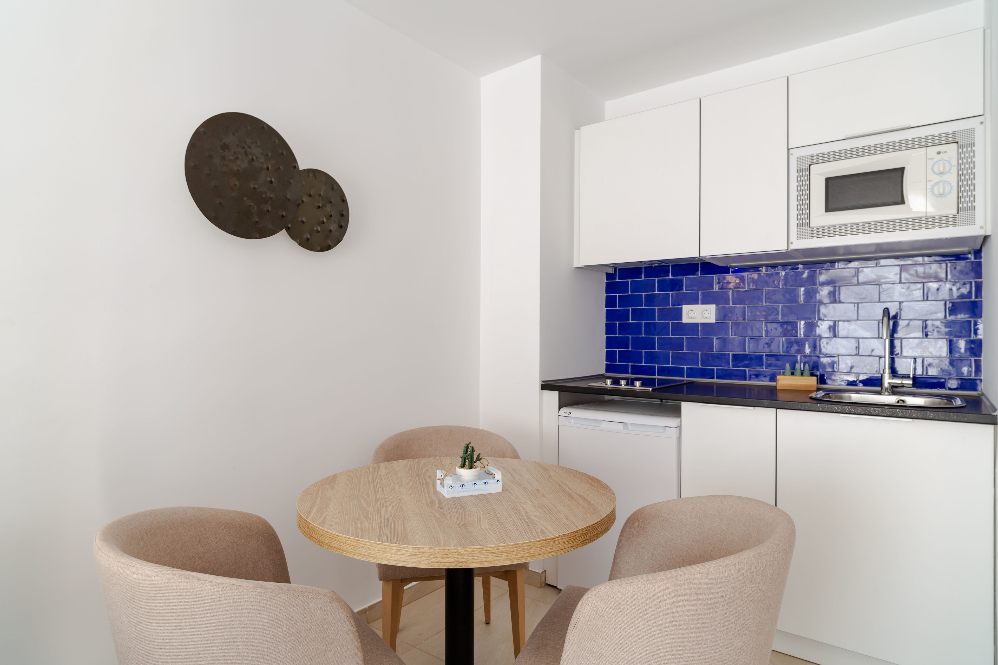 Een compacte keuken met witte kasten, een blauw betegelde achterwand, een magnetron en een ronde houten tafel met stoelen.
