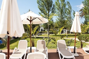 Una terraza con sillas reclinables blancas y sombrillas, rodeada de vegetación y un cielo despejado.