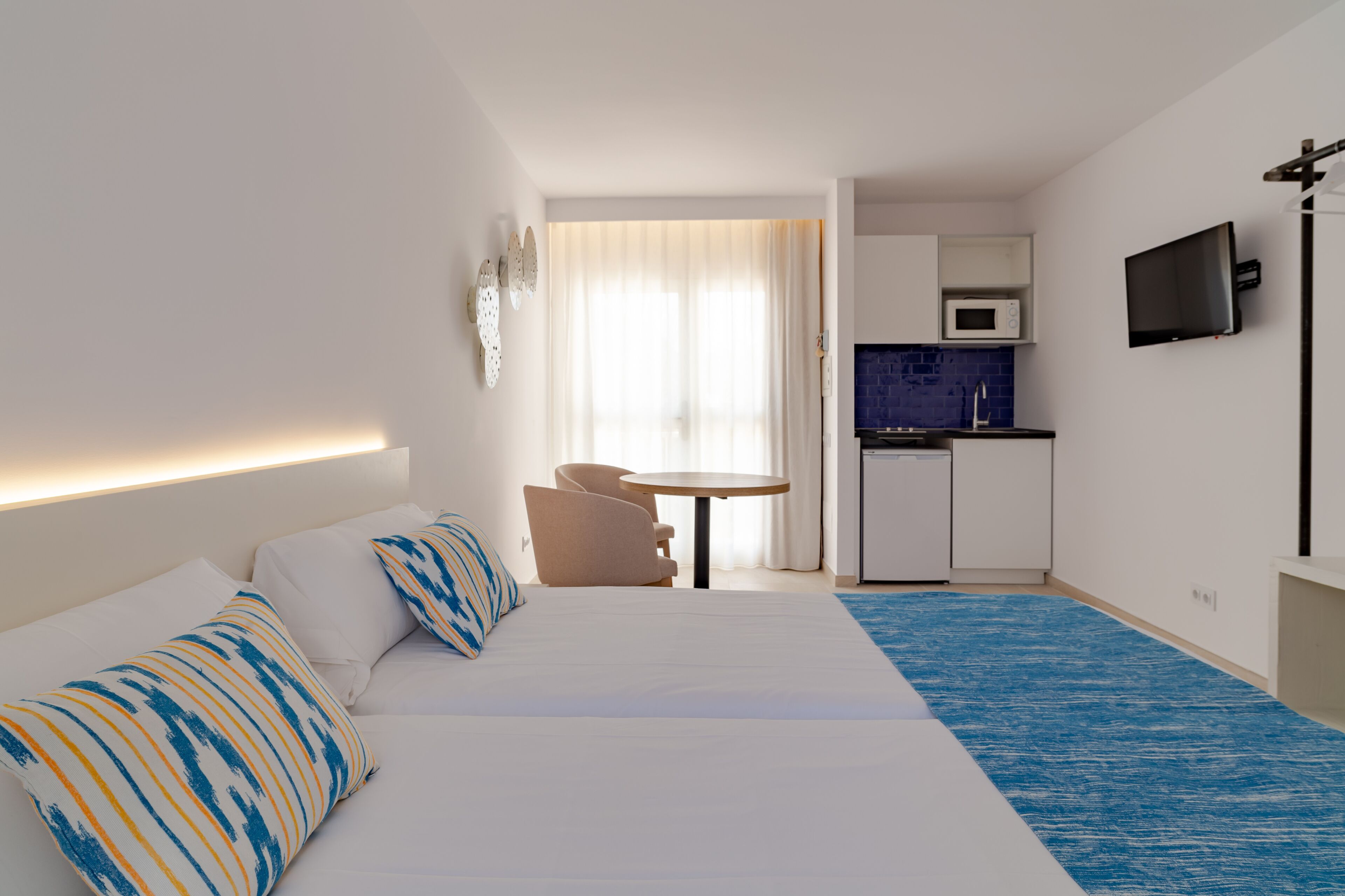 Habitació d'hotel amb llit, una cuineta petita, televisió i finestra amb cortines.