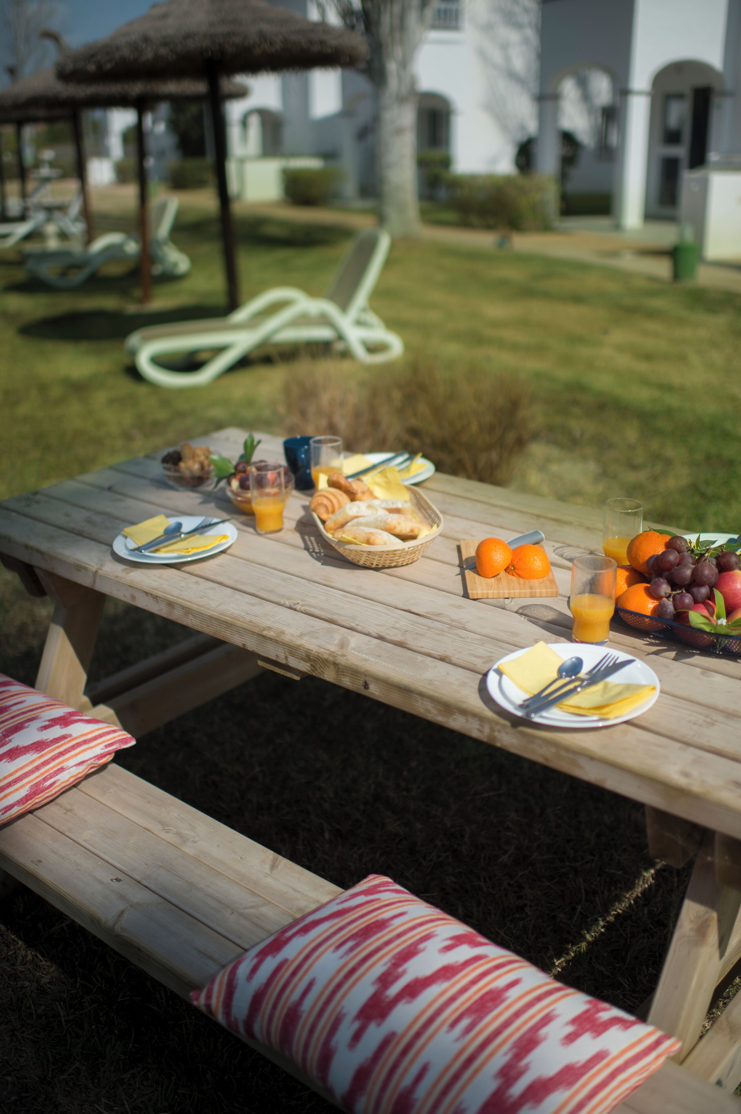 Une table de pique-nique avec de la nourriture et des boissons, un parasol en chaume et un banc agrémenté de coussins à motifs.