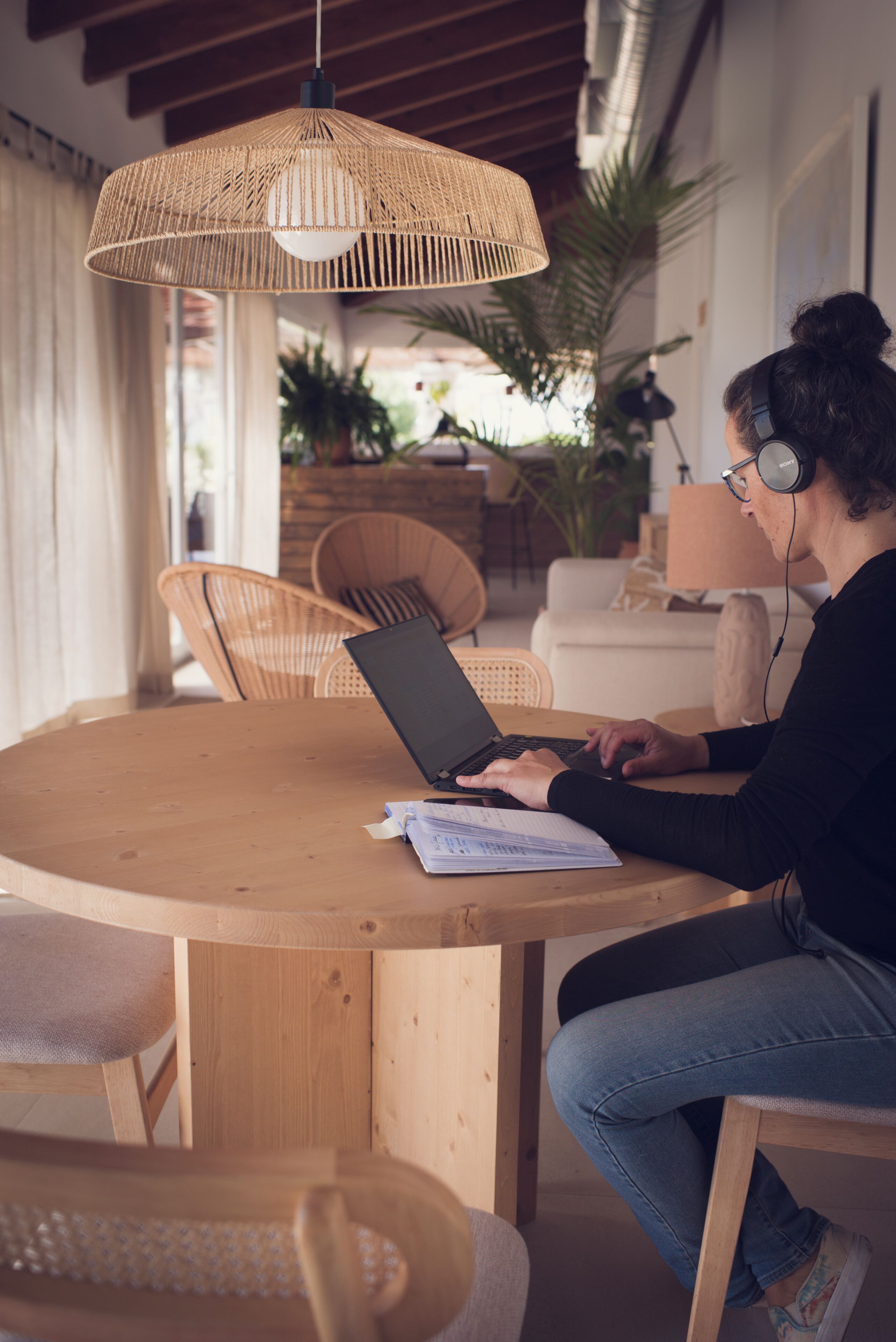 Une personne utilise un ordinateur portable à une table en bois, sous une suspension décorative en fibres tressées. Des chaises en osier et une plante en pot l’entourent.