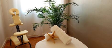 A room with a massage chair, a large potted plant, and a neatly folded towel on a white table.