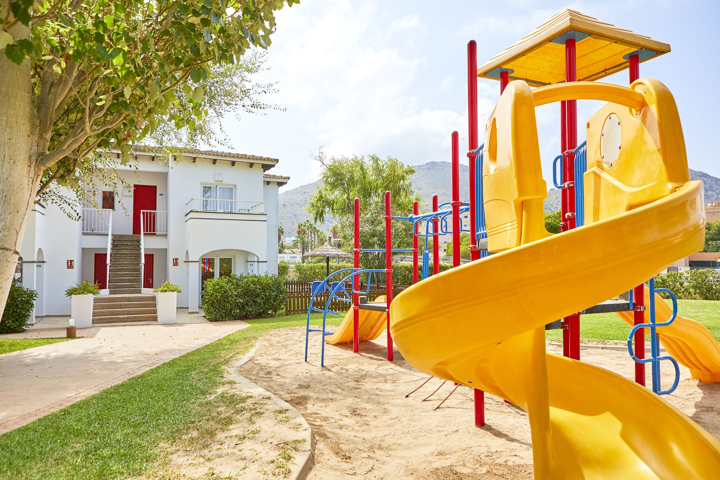 Un parque infantil con un tobogán amarillo y estructuras de escalada, un edificio con puerta roja y una escalera que lleva a un balcón.
