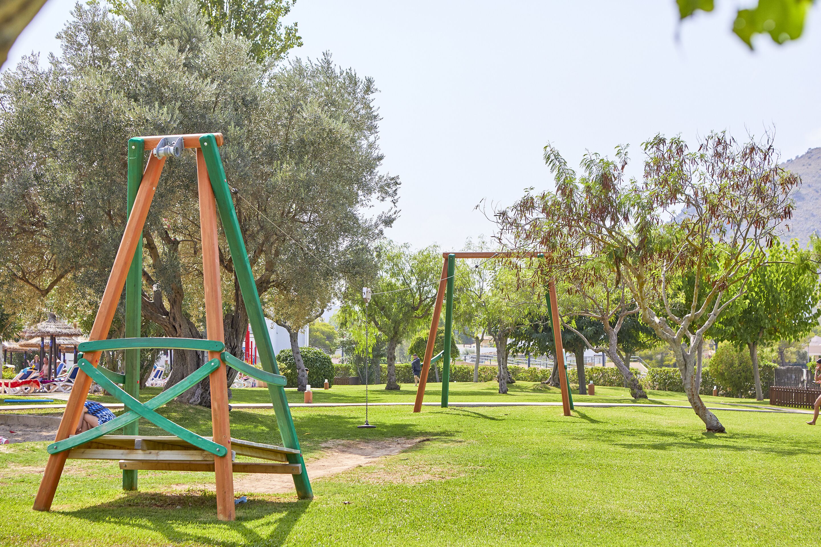 A playground with swings and a slide, surrounded by grass and trees.