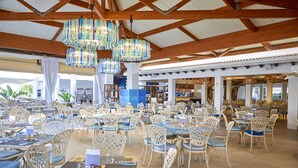 Un restaurante espacioso con sillas y mesas de mimbre blanco, grandes candelabros colgantes y un techo alto con vigas de madera.